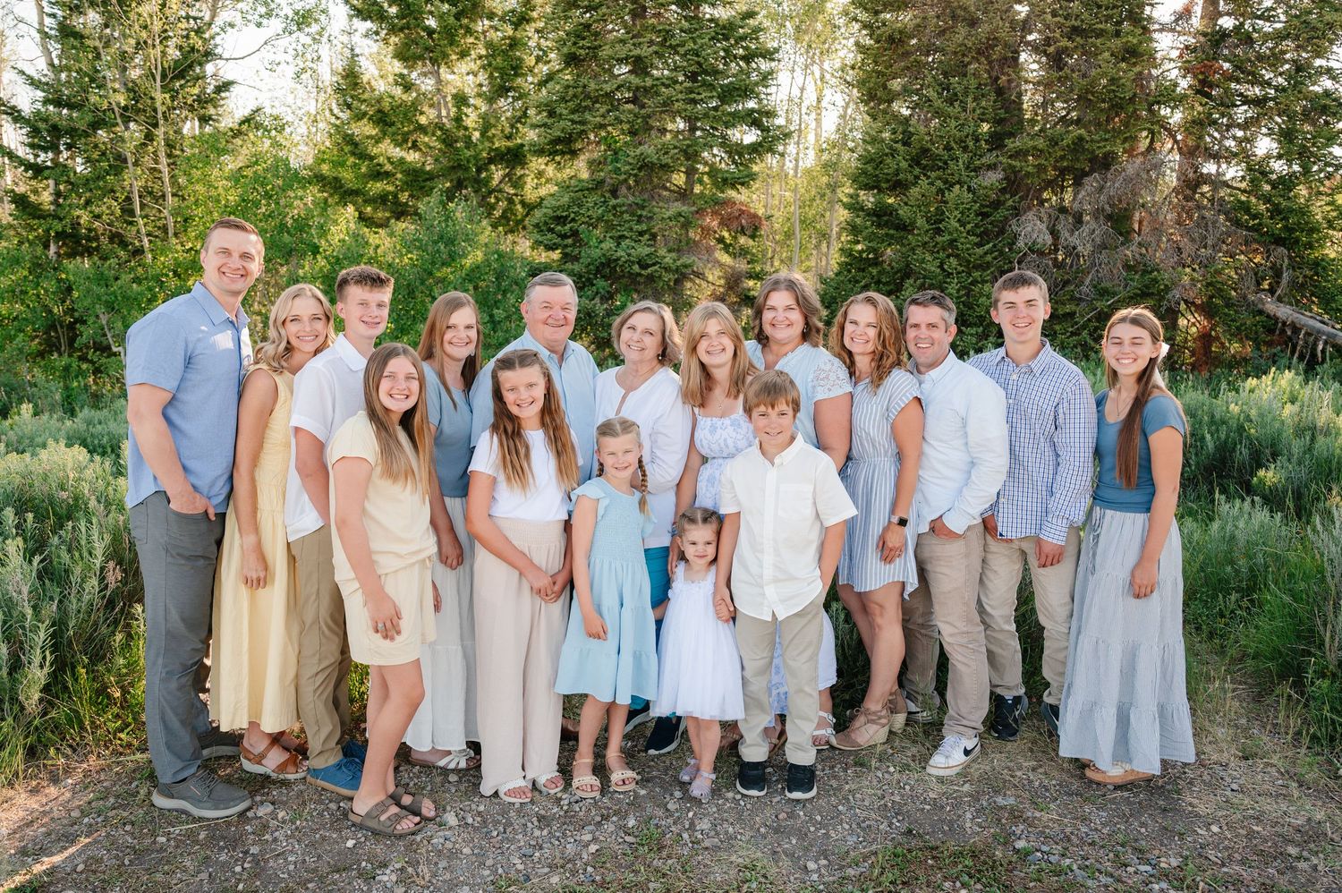 Large family group portrait outdoors among pine trees, dressed in coordinating white and neutral tones.