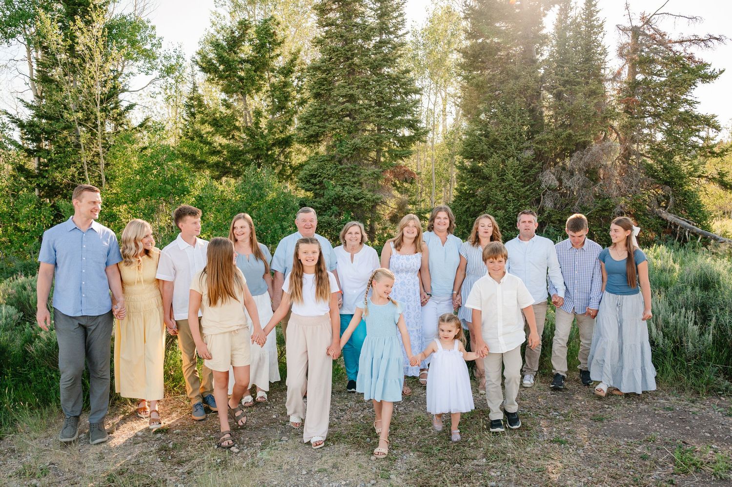 Large family group photo outdoors in nature, wearing coordinated pastel outfits, holding hands and smiling among pine trees.