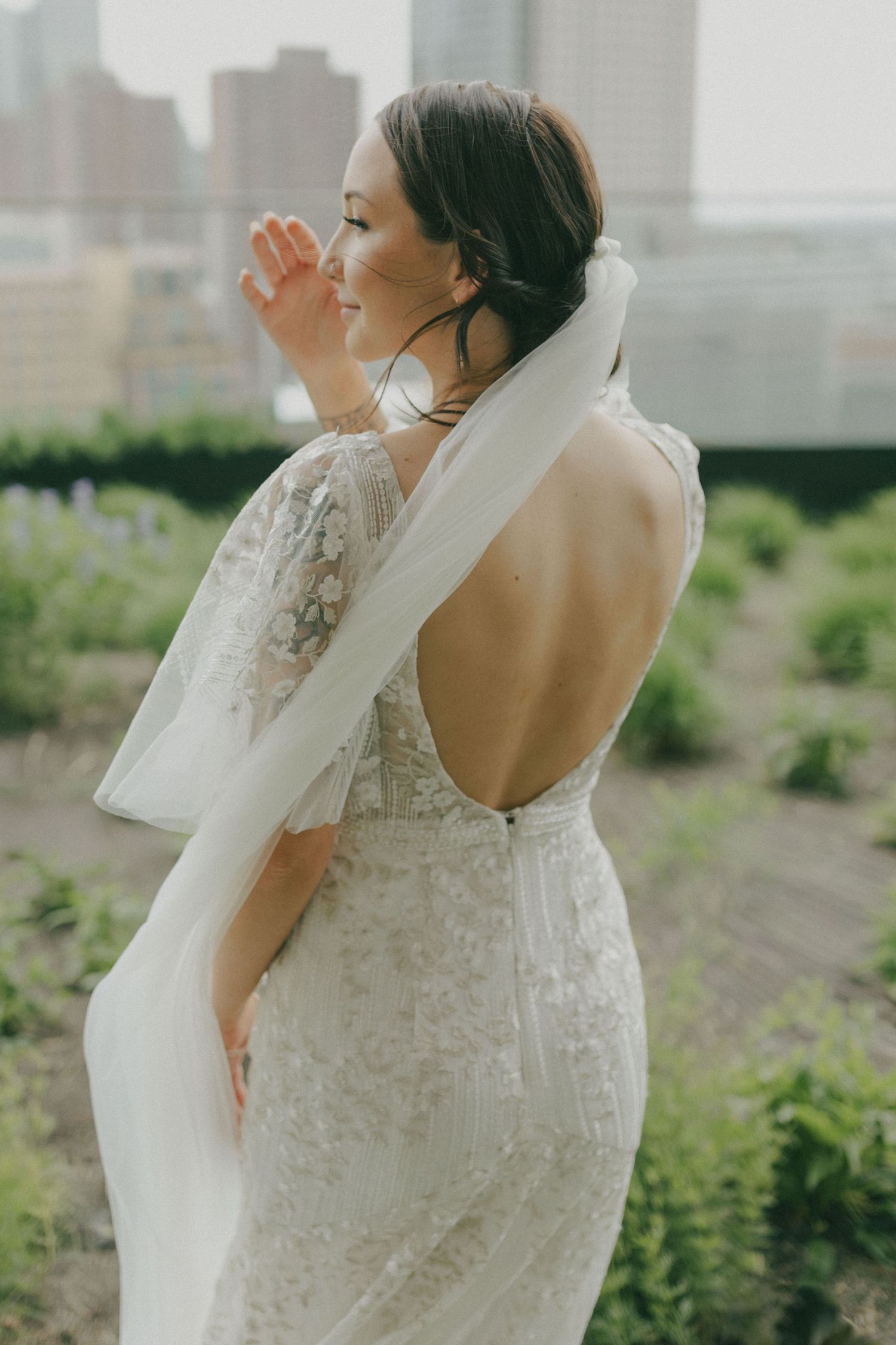 Bride in backless lace wedding gown with flowing veil standing on rooftop garden with city skyline backdrop.
