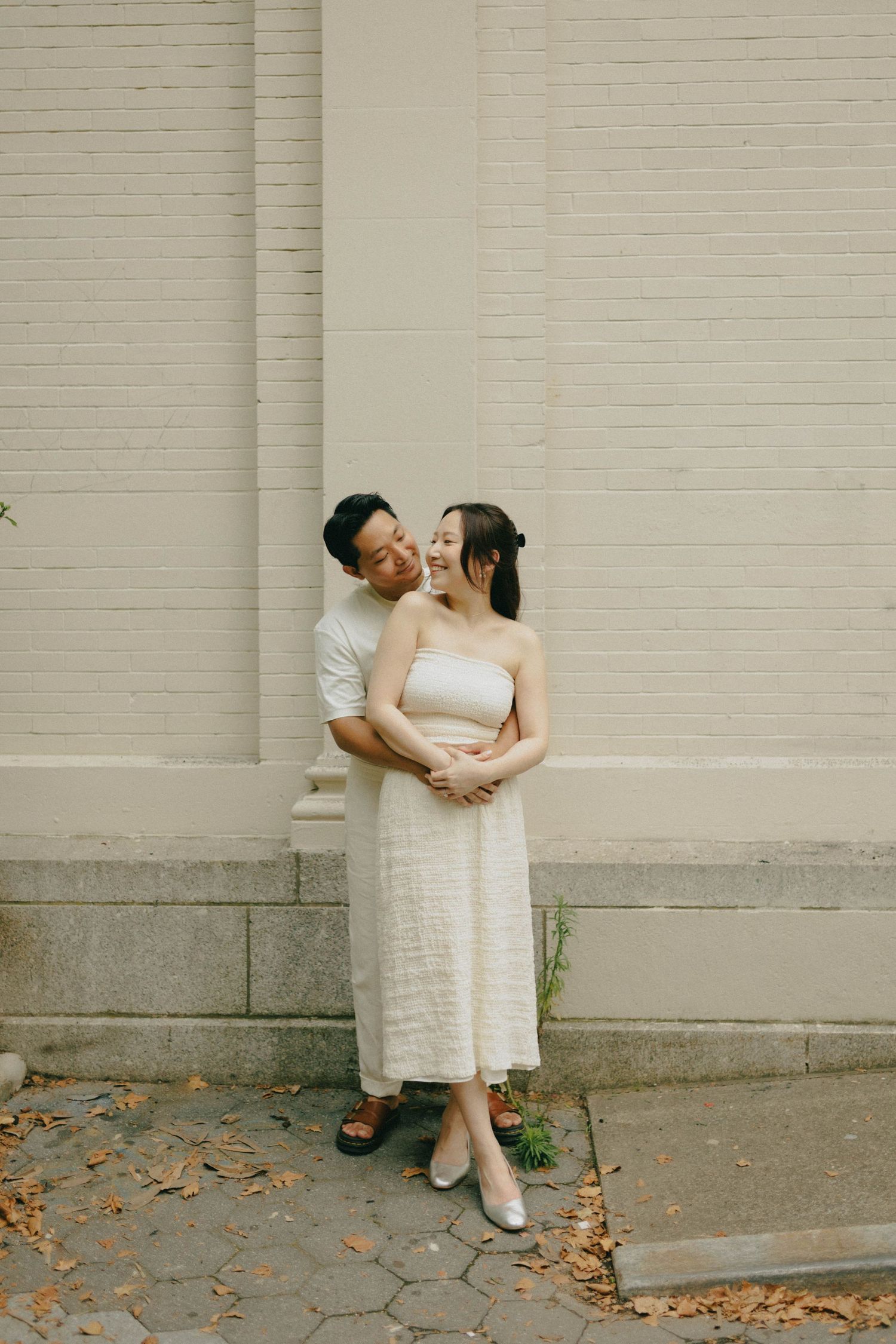 Asian couple embracing and smiling together against a cream brick wall surrounded by fallen autumn leaves.