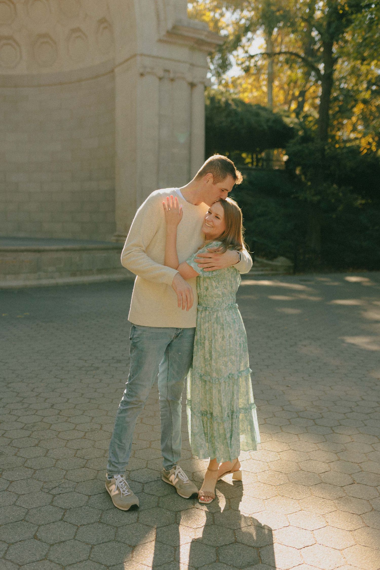 Couple embracing in golden sunset light near ornate stone archway, woman wearing floral green midi dress.