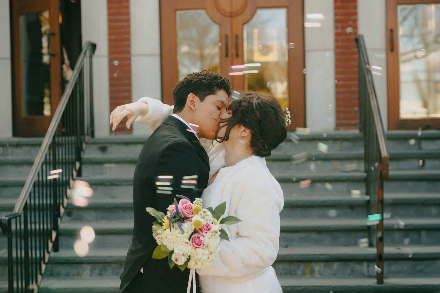 Newlyweds sharing a kiss on church steps as bubbles float around them, bride holding pink bouquet.