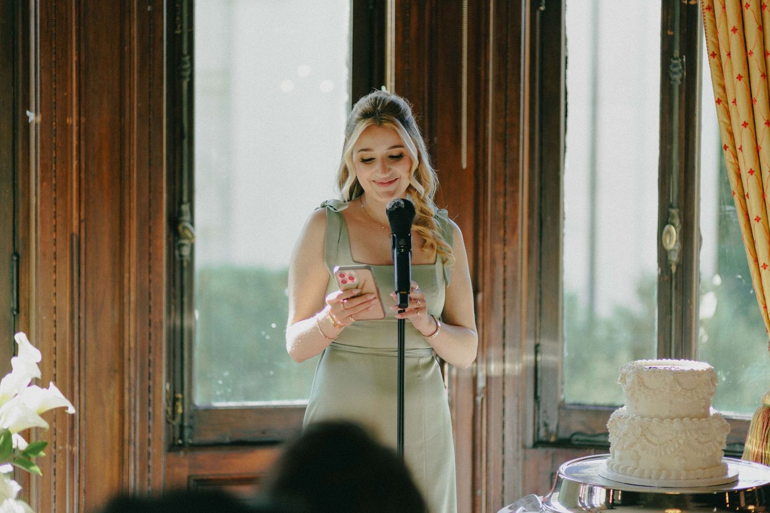 Elegant bridesmaid in sage green gown delivering speech with microphone and phone at wedding reception.