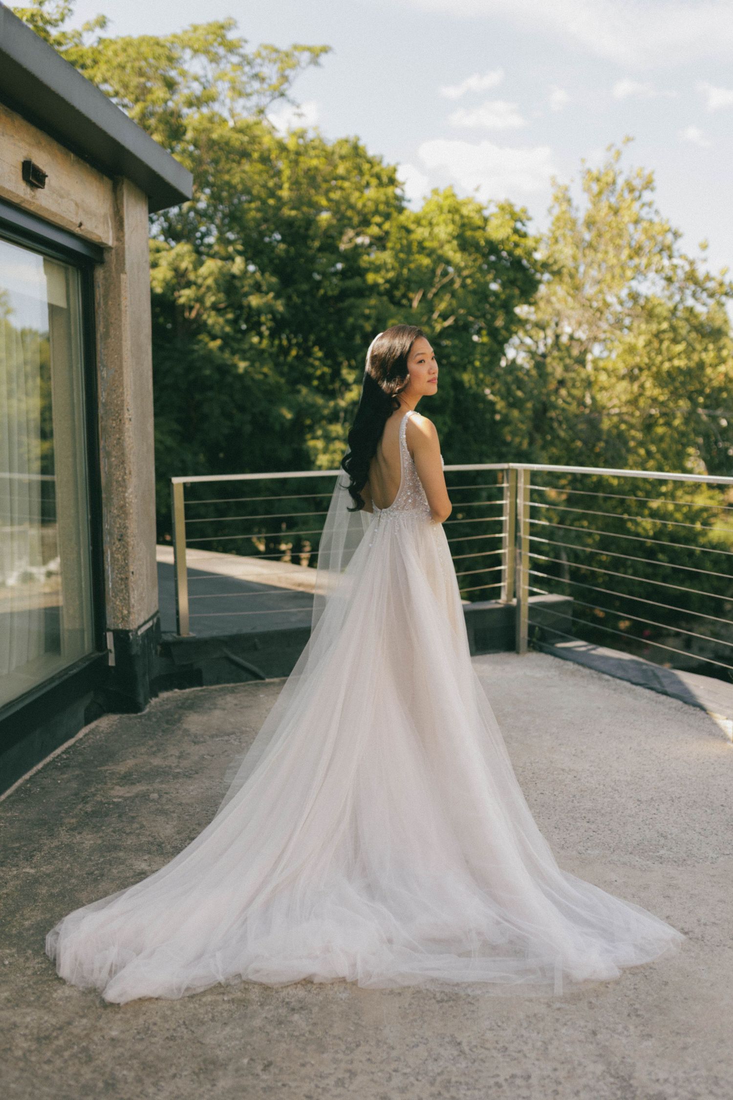 Elegant bride in flowing white tulle gown standing on balcony surrounded by lush green trees.