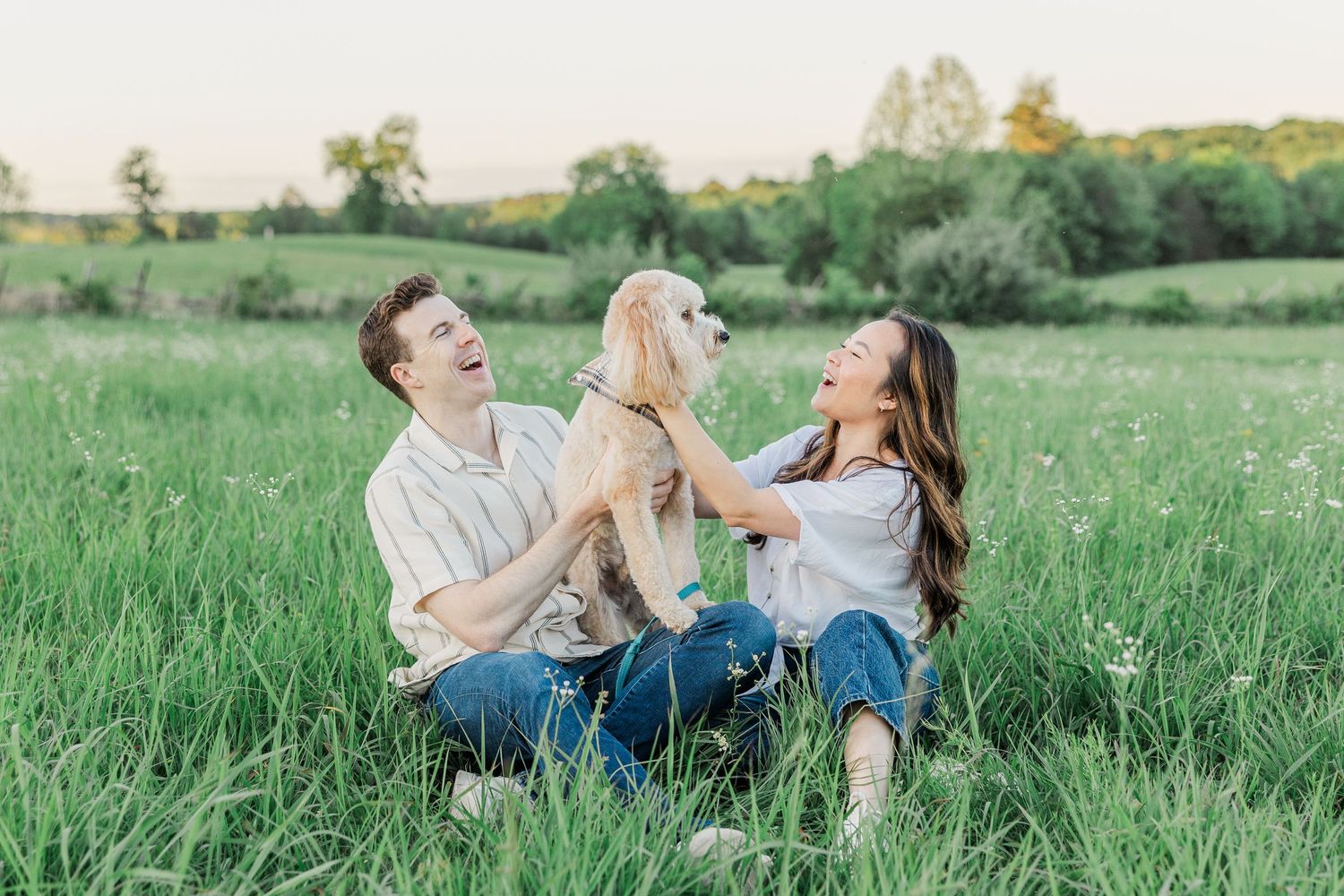 Couple sitting in green field laughing and playing with golden retriever dog at sunset.