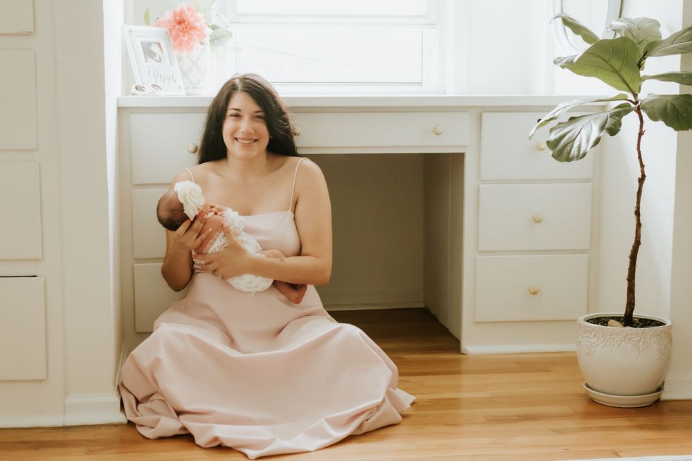 A smiling mother in a blush pink dress sits on hardwood floor cradling her newborn baby near white drawers.