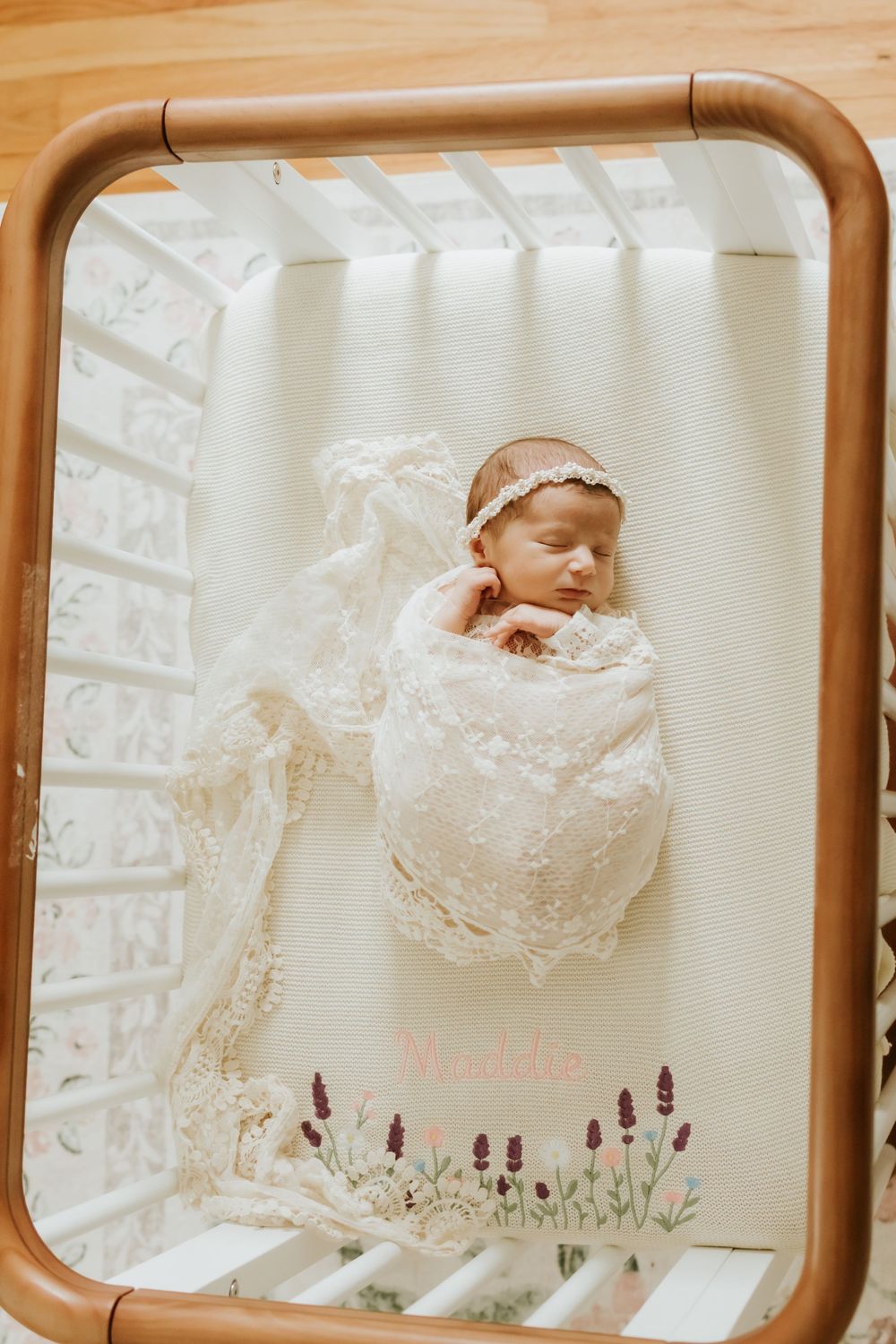 Sleeping newborn baby swaddled in lace wrap with floral headband lying in a wooden crib with embroidered bedding.