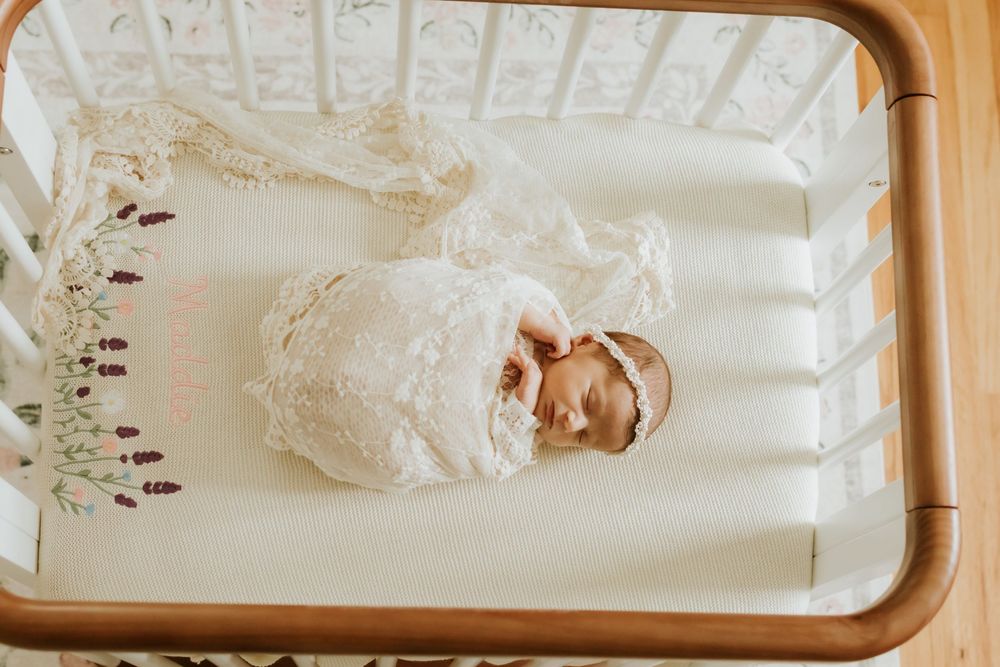 Newborn baby sleeping peacefully in a white crib, dressed in a delicate lace outfit with a floral headband.