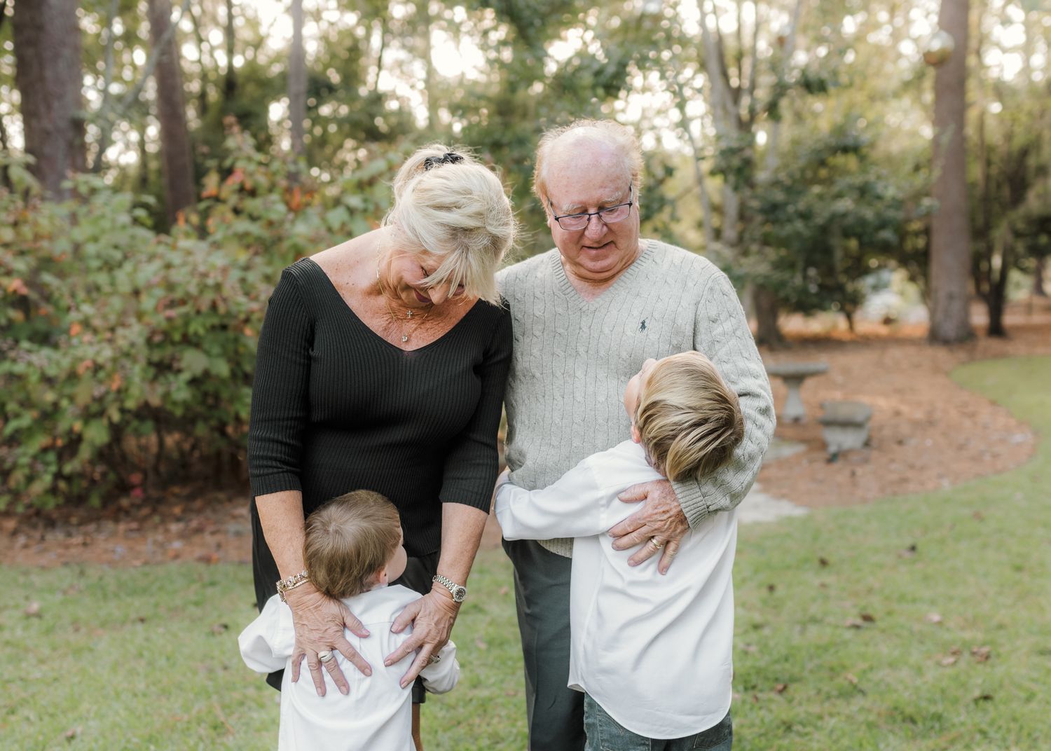 Grandparents lovingly embracing two young grandchildren outdoors in a lush green park setting.