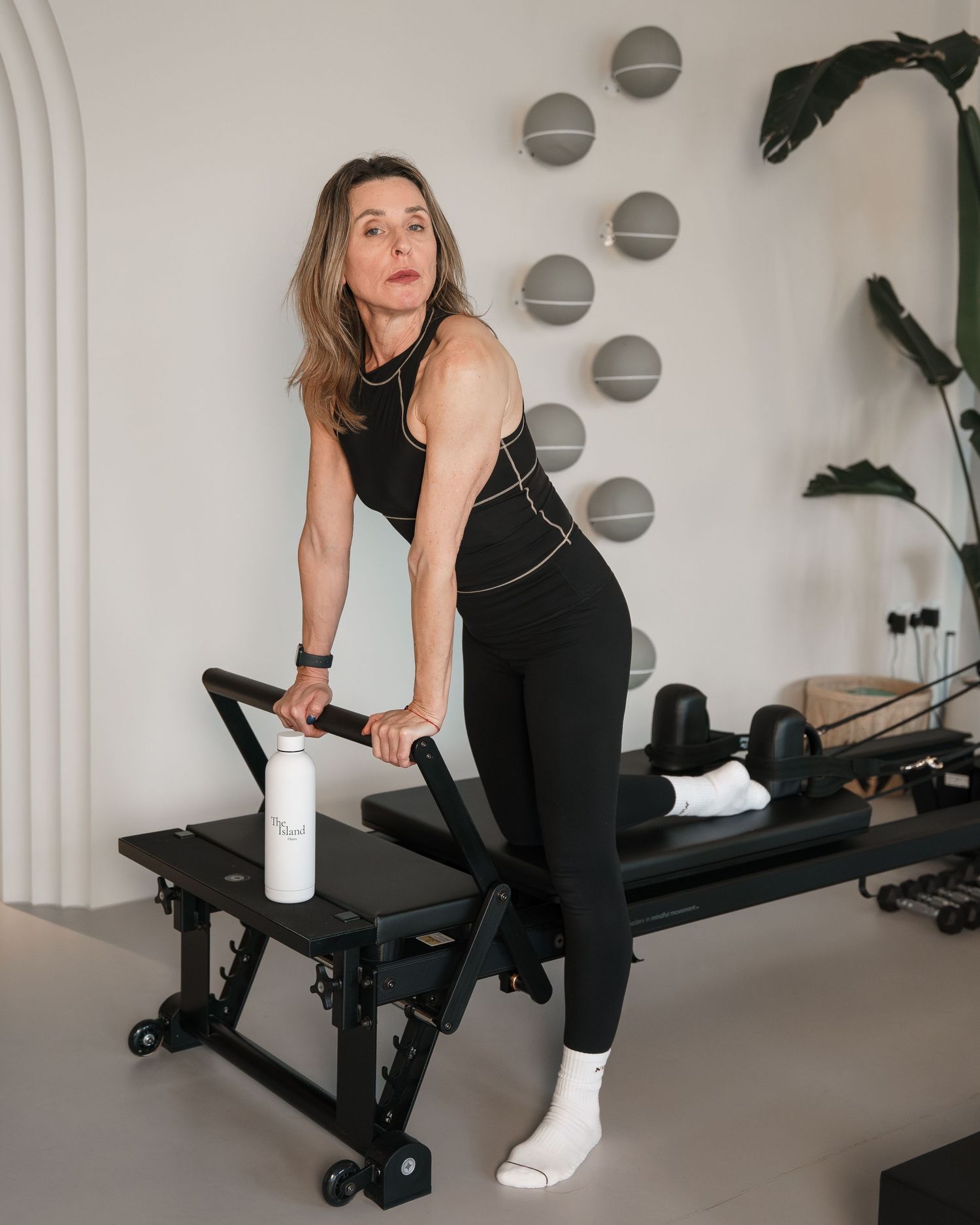 Woman in black athletic wear doing Pilates on a reformer machine in a modern white studio.