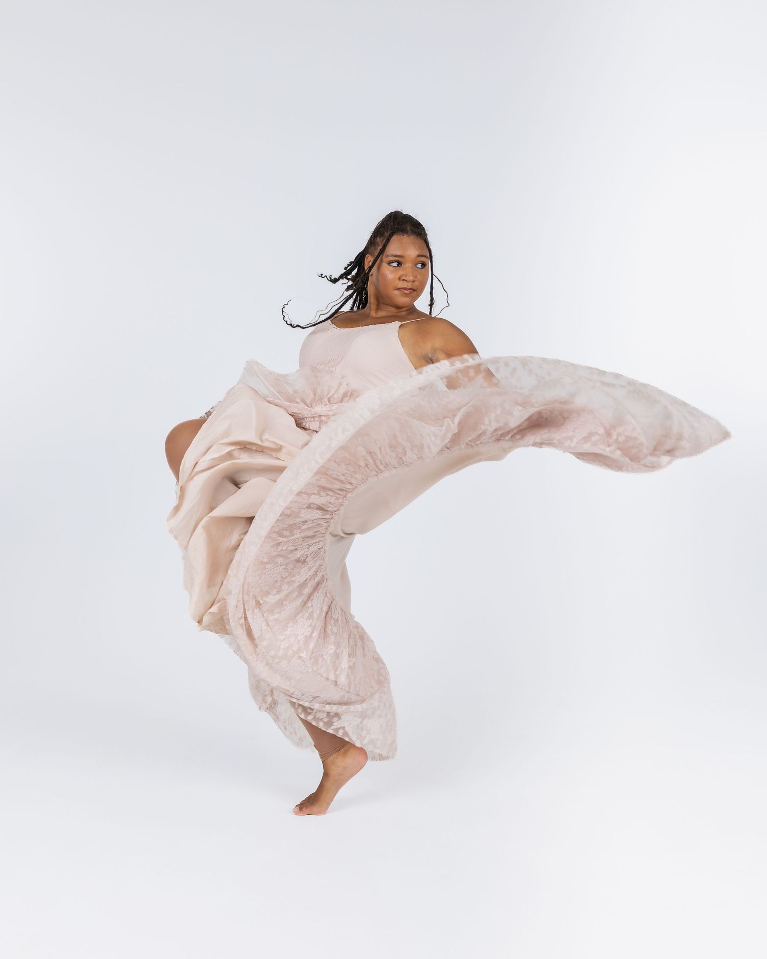 A dancer in a flowing blush pink gown leaps gracefully against a white studio background.