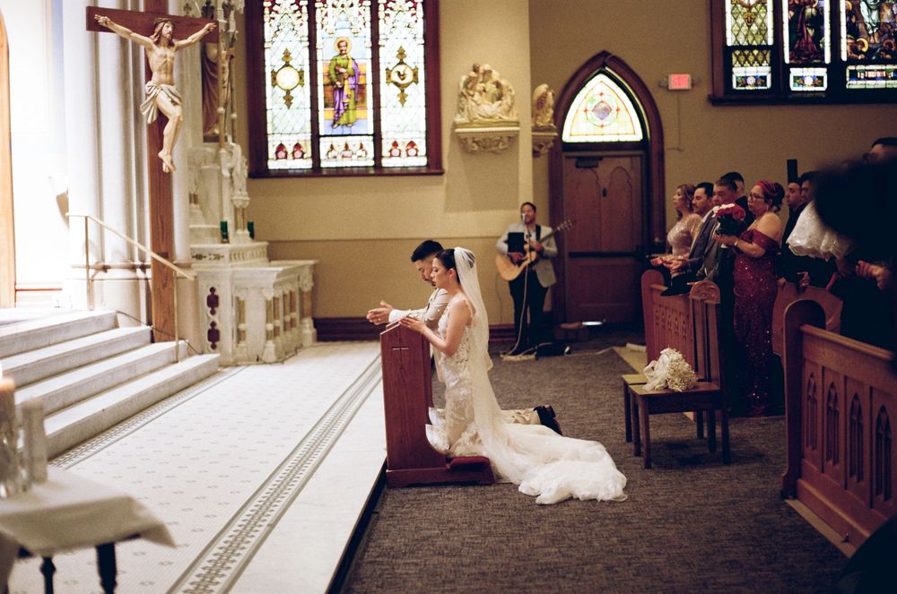Bride in white gown kneeling at church altar during Catholic wedding ceremony with guests seated in pews.