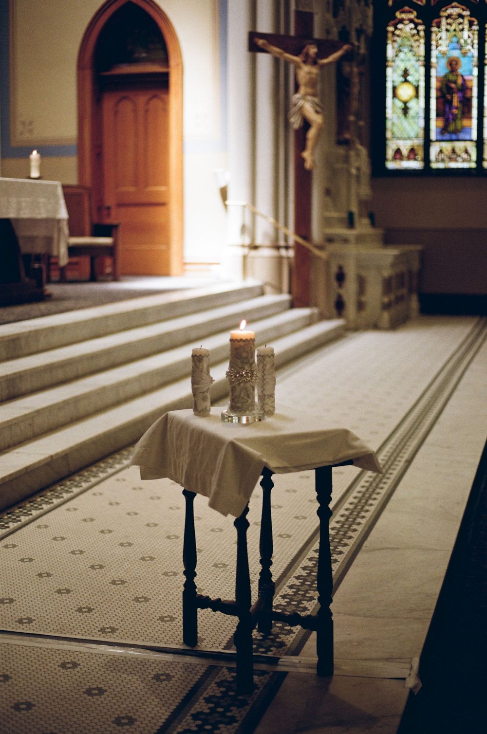 Lit candles on a small altar table inside a Catholic church with crucifix and stained glass windows.