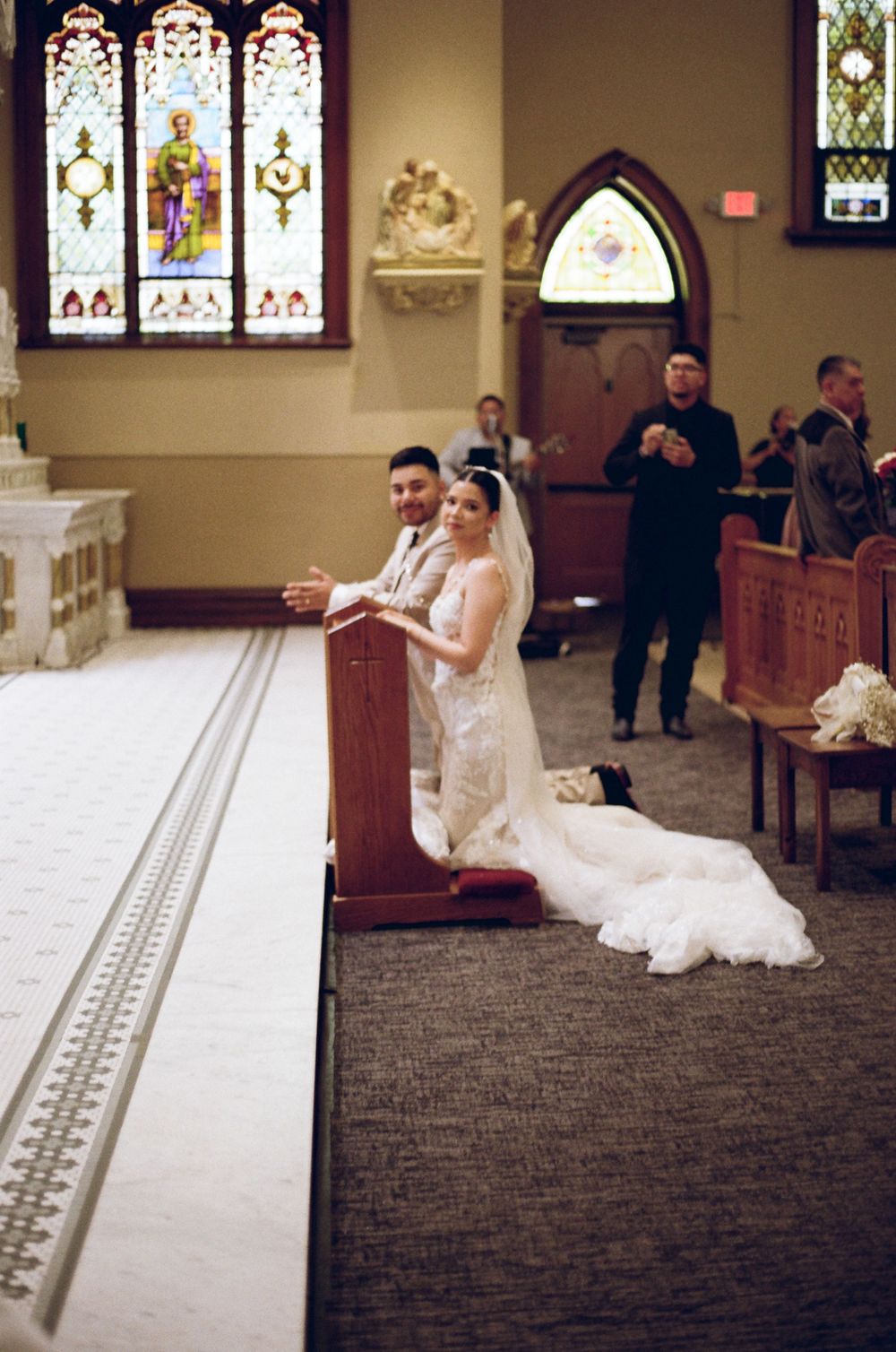 Bride and groom kneeling at church altar during wedding ceremony with stained glass windows in background.