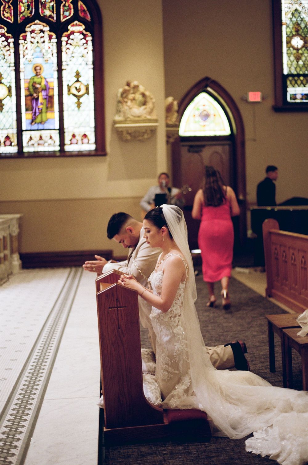 Bride and groom kneeling in prayer at church pew during wedding ceremony with stained glass windows.