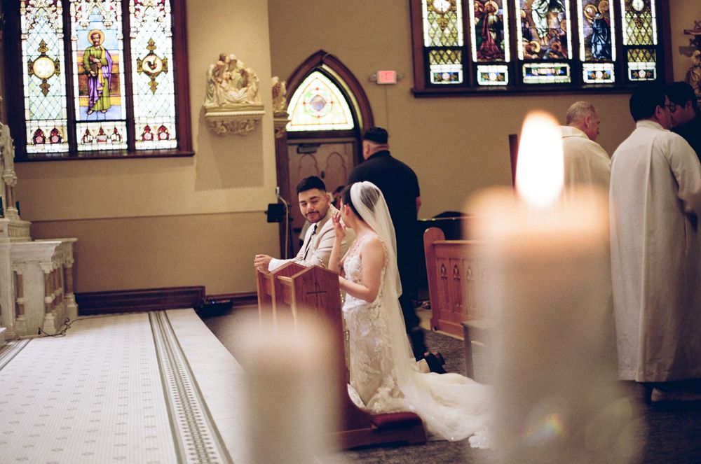 Bride and groom kneeling at altar during church wedding ceremony with stained glass windows and candles.