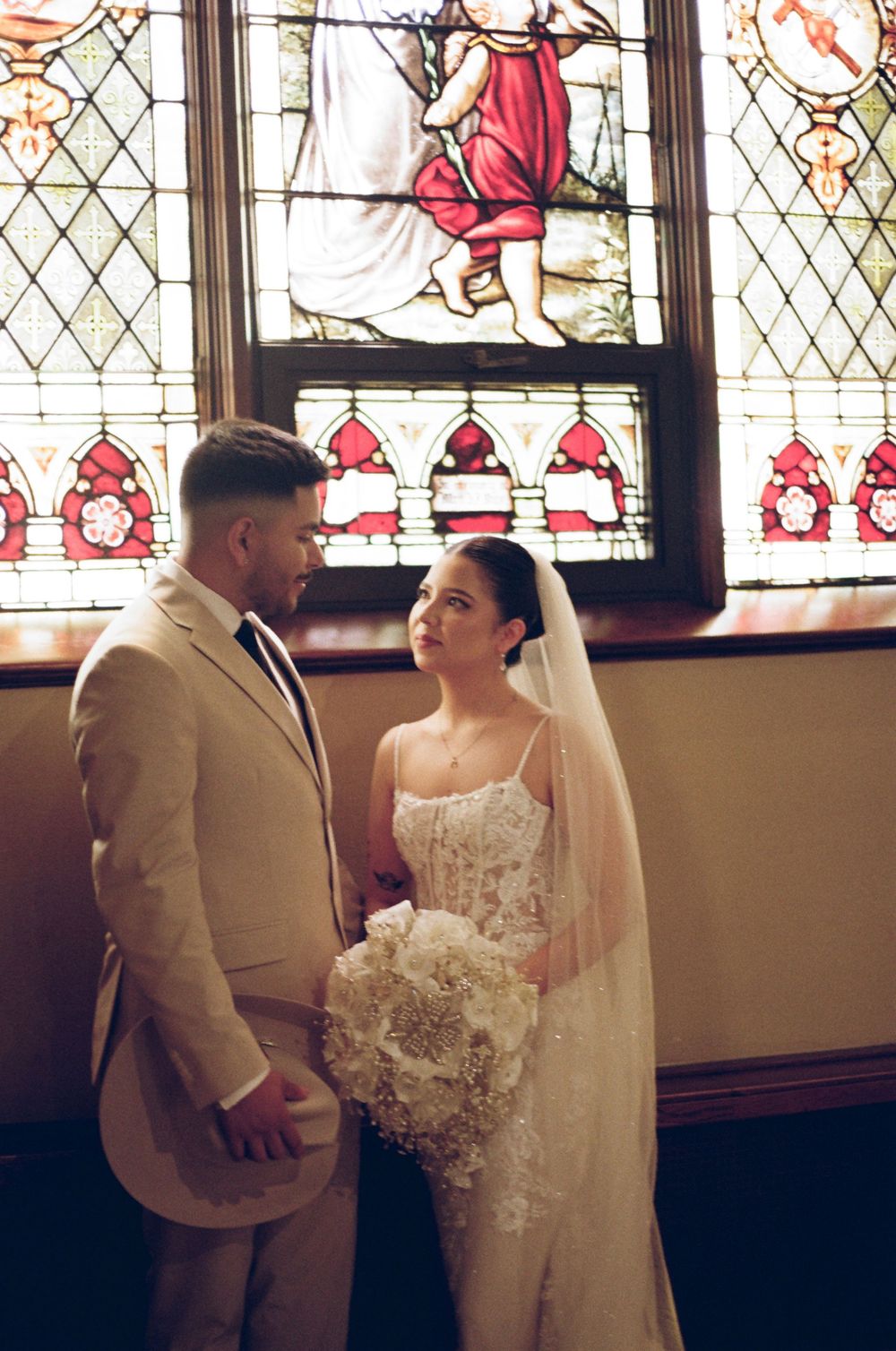 Bride and groom share a romantic moment before a stunning stained glass church window, bride holding white floral bouquet.