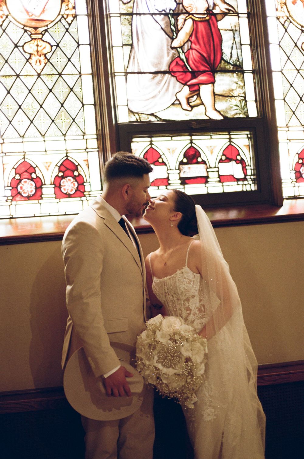 Bride and groom share a kiss in front of stunning stained glass window, bride holding white bouquet.