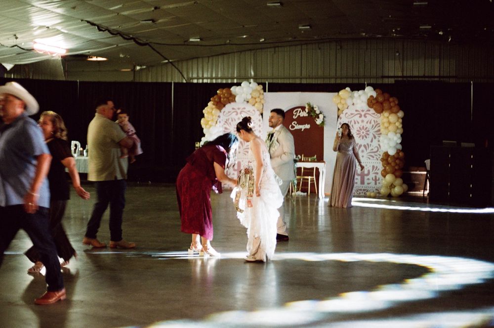 Bride in white gown stands on dance floor at wedding reception with balloon arch backdrop in decorated event hall.