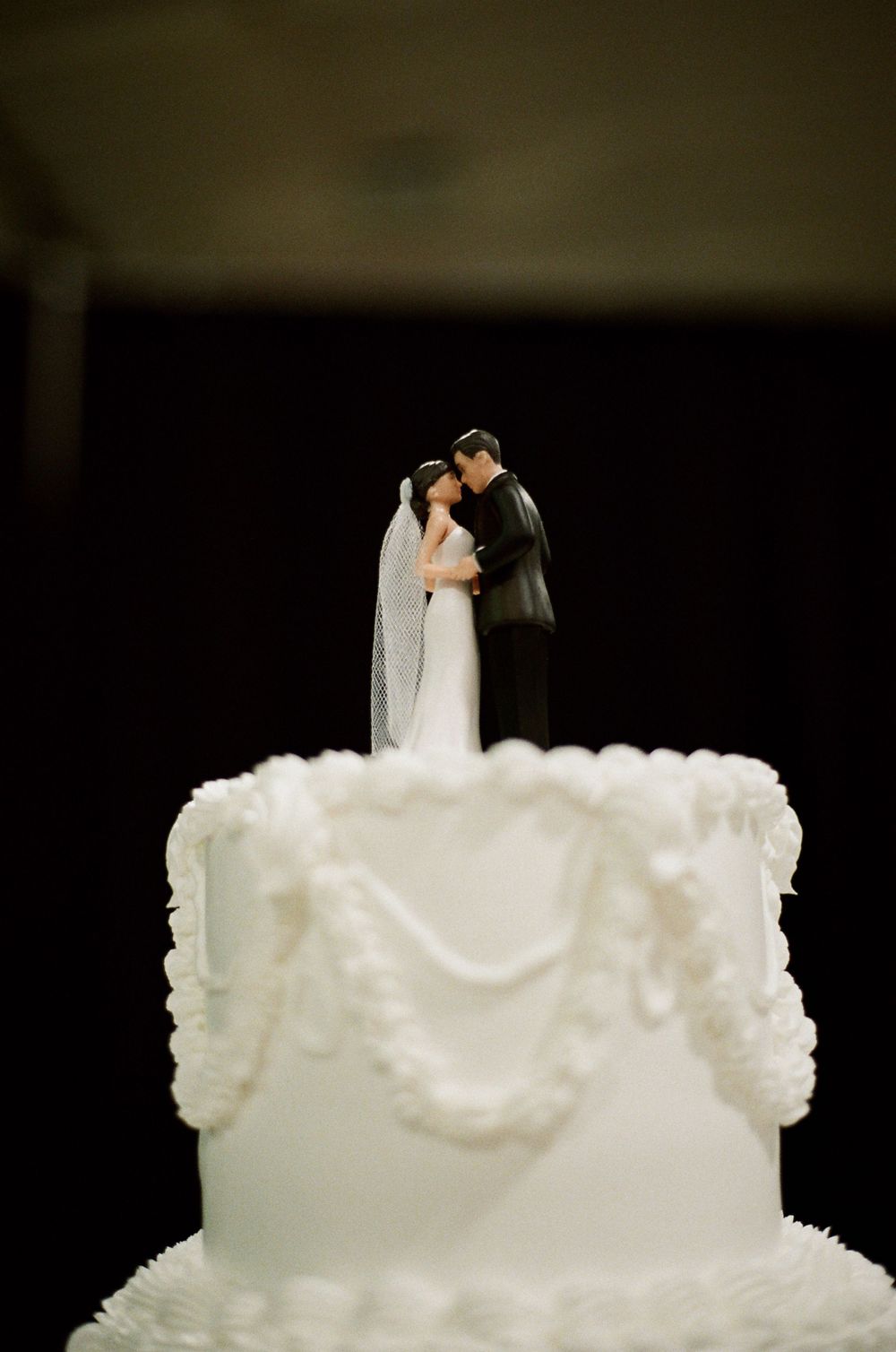 White wedding cake with elegant frosting and classic bride and groom figurine topper kissing on dark background.