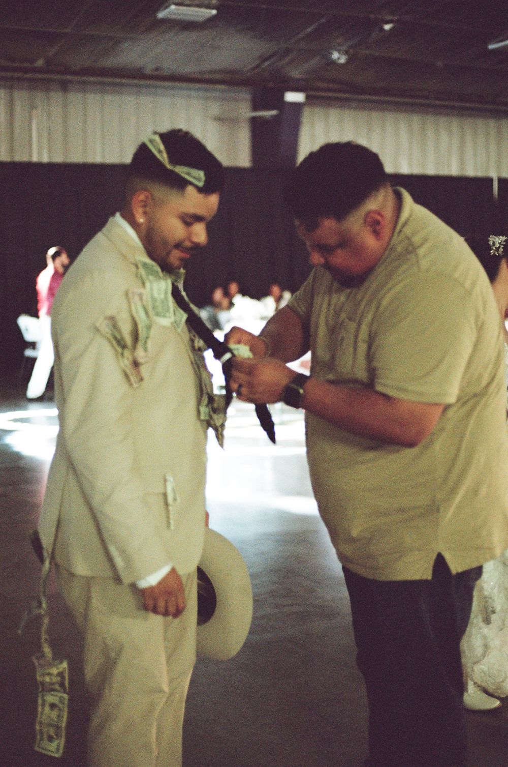 A groom in a beige suit receives help adjusting his tie from a man in a light green shirt at a wedding venue.