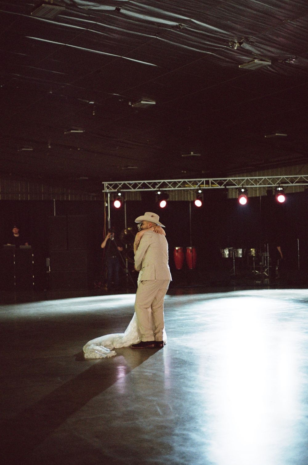 A performer in a white suit and hat stands alone on a dimly lit dance floor with stage lights in the background.