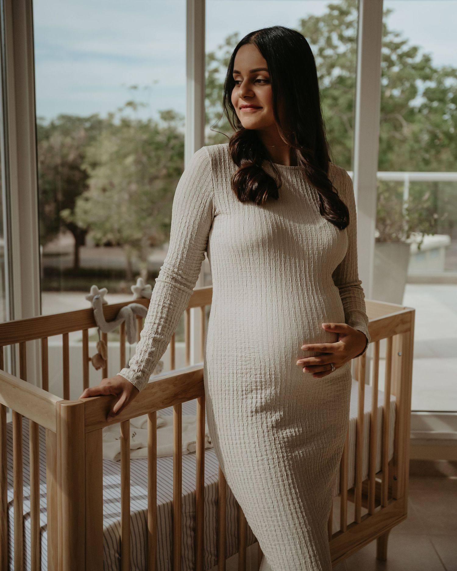 Pregnant woman in cream ribbed maxi dress standing beside wooden baby crib, smiling near large windows.