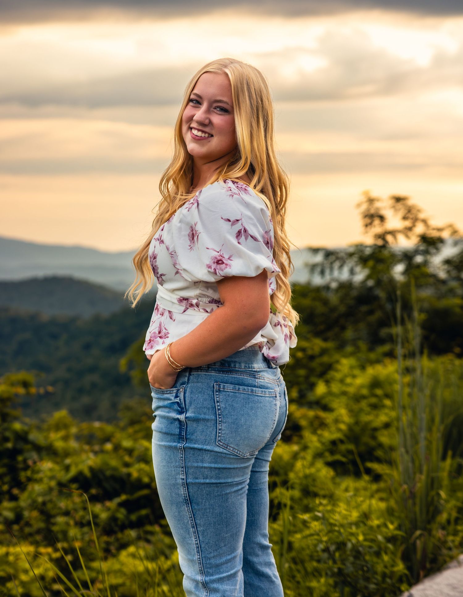Young blonde woman in white lace dress sitting on green hillside holding a book with mountain views behind her.