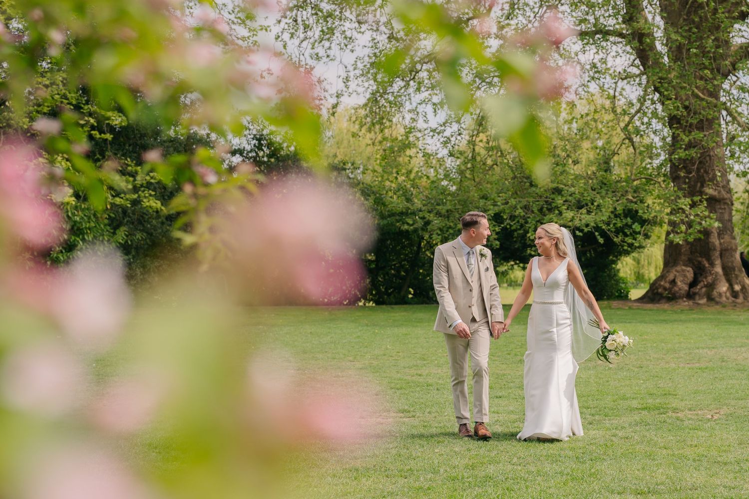 Bride and groom walking hand in hand through lush green garden framed by pink blossoms on sunny wedding day.