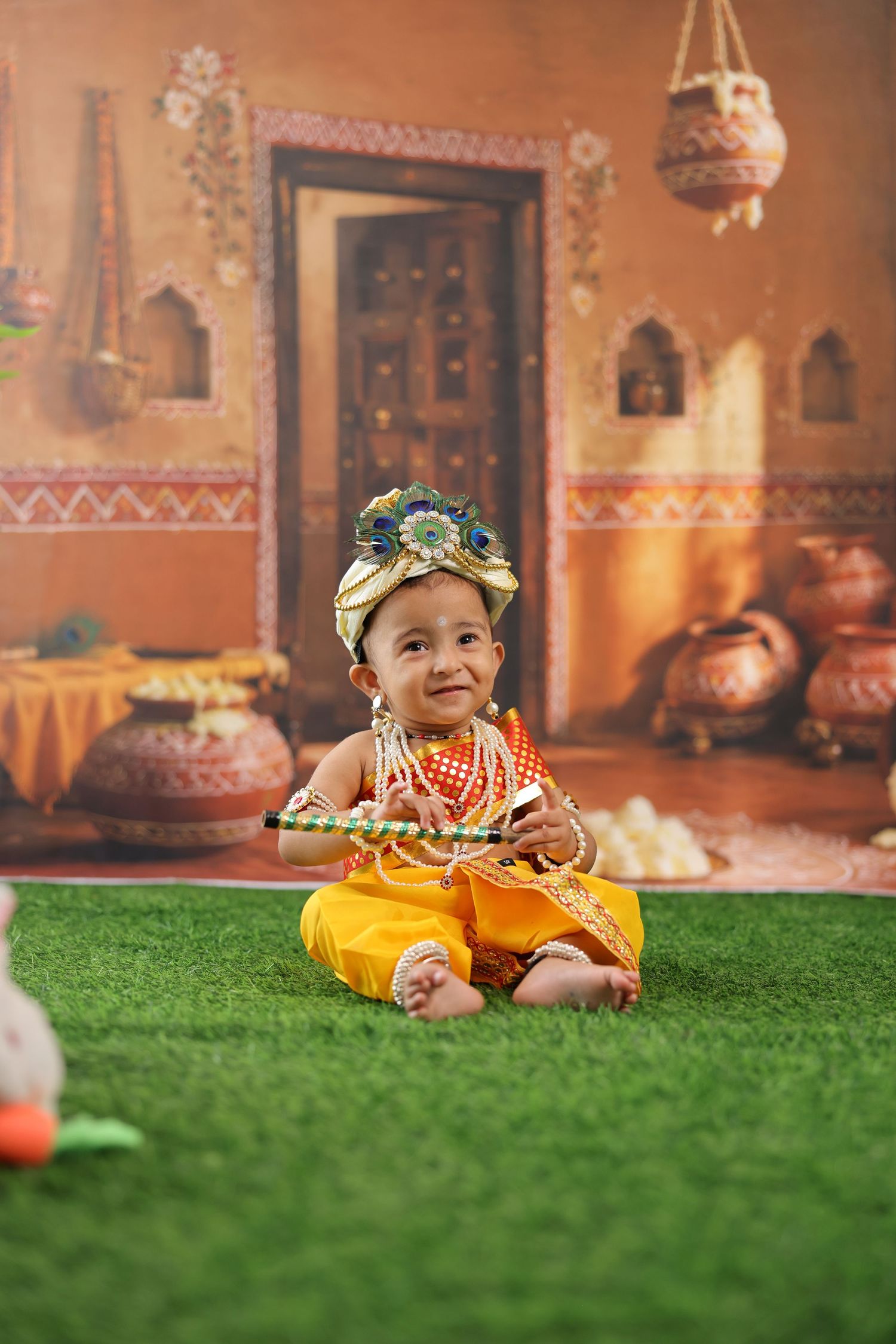 A baby dressed as Lord Krishna in yellow outfit and floral crown sits on grass with traditional Indian backdrop.