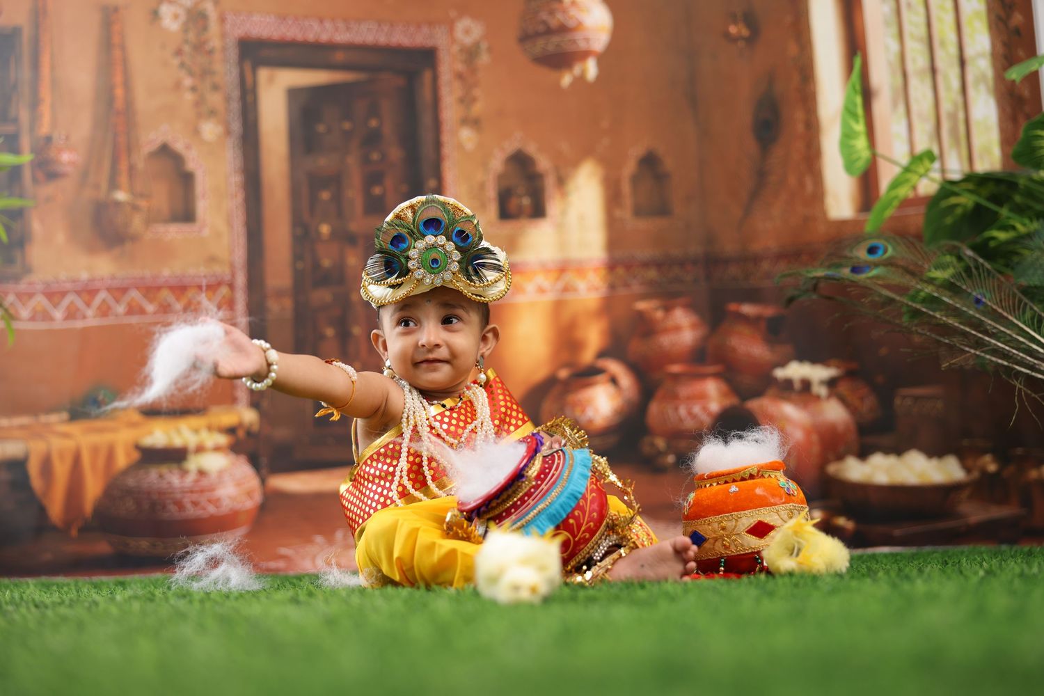 Baby dressed as Lord Krishna in colorful traditional costume with peacock feather crown, sitting on green grass.