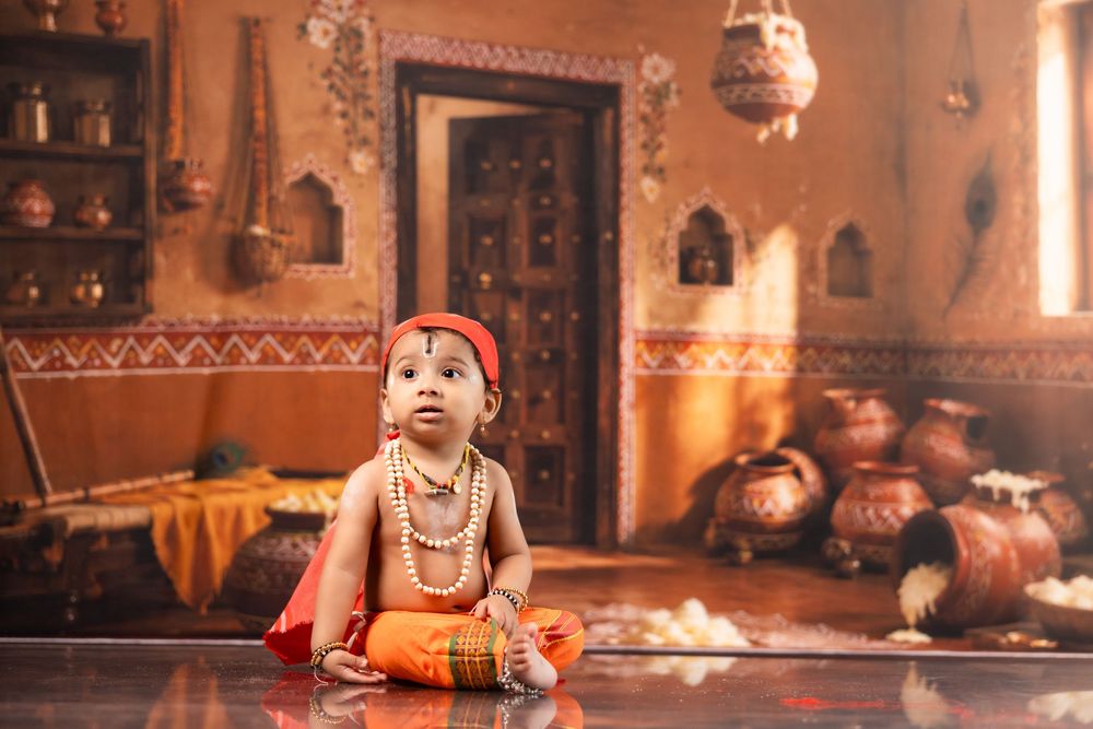Baby dressed as Lord Krishna in traditional Indian attire, sitting in a decorative ethnic home setting.