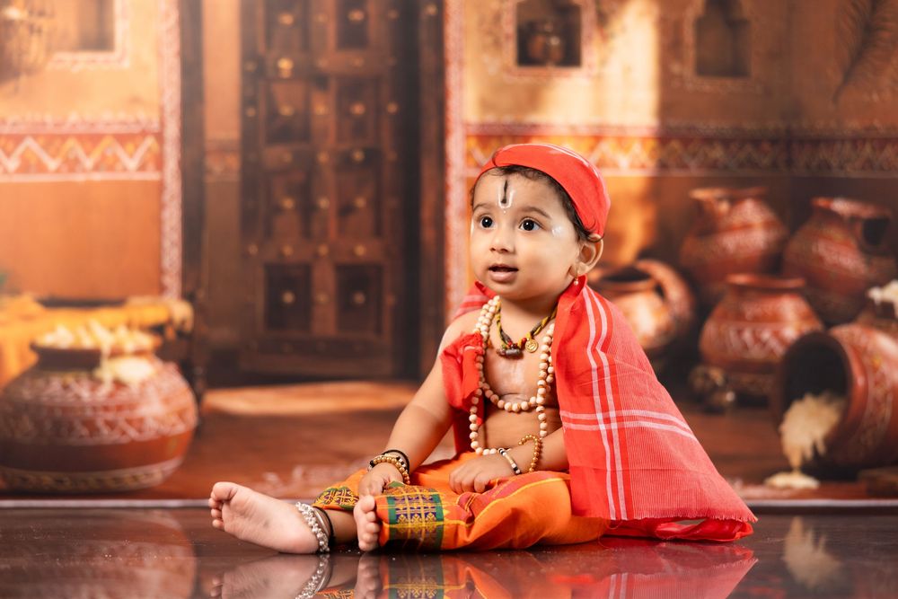 Adorable baby dressed in traditional Indian attire with orange outfit, jewelry, and red cap, seated on floor.