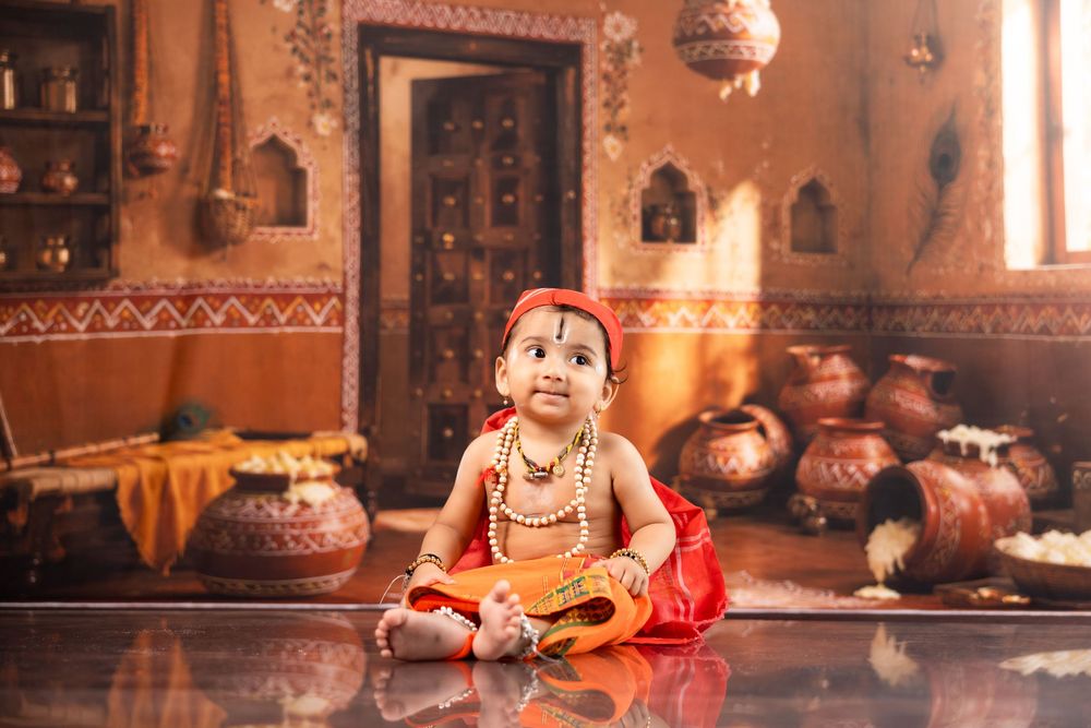 A baby dressed as Lord Krishna in traditional red outfit with jewelry, sitting in a decorated Indian temple setting.