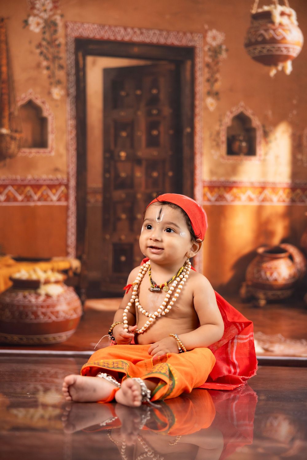 A baby dressed as Lord Krishna in traditional orange attire with beads and red cap sits on a reflective floor in a decorated Indian setting.