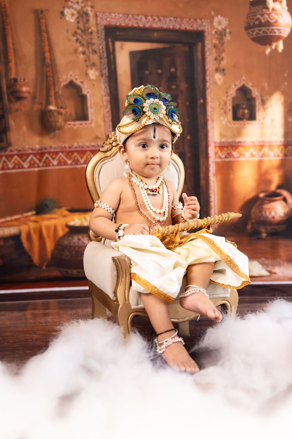 Baby dressed as Lord Krishna in traditional attire with peacock crown, beads, and flute, seated on ornate chair.