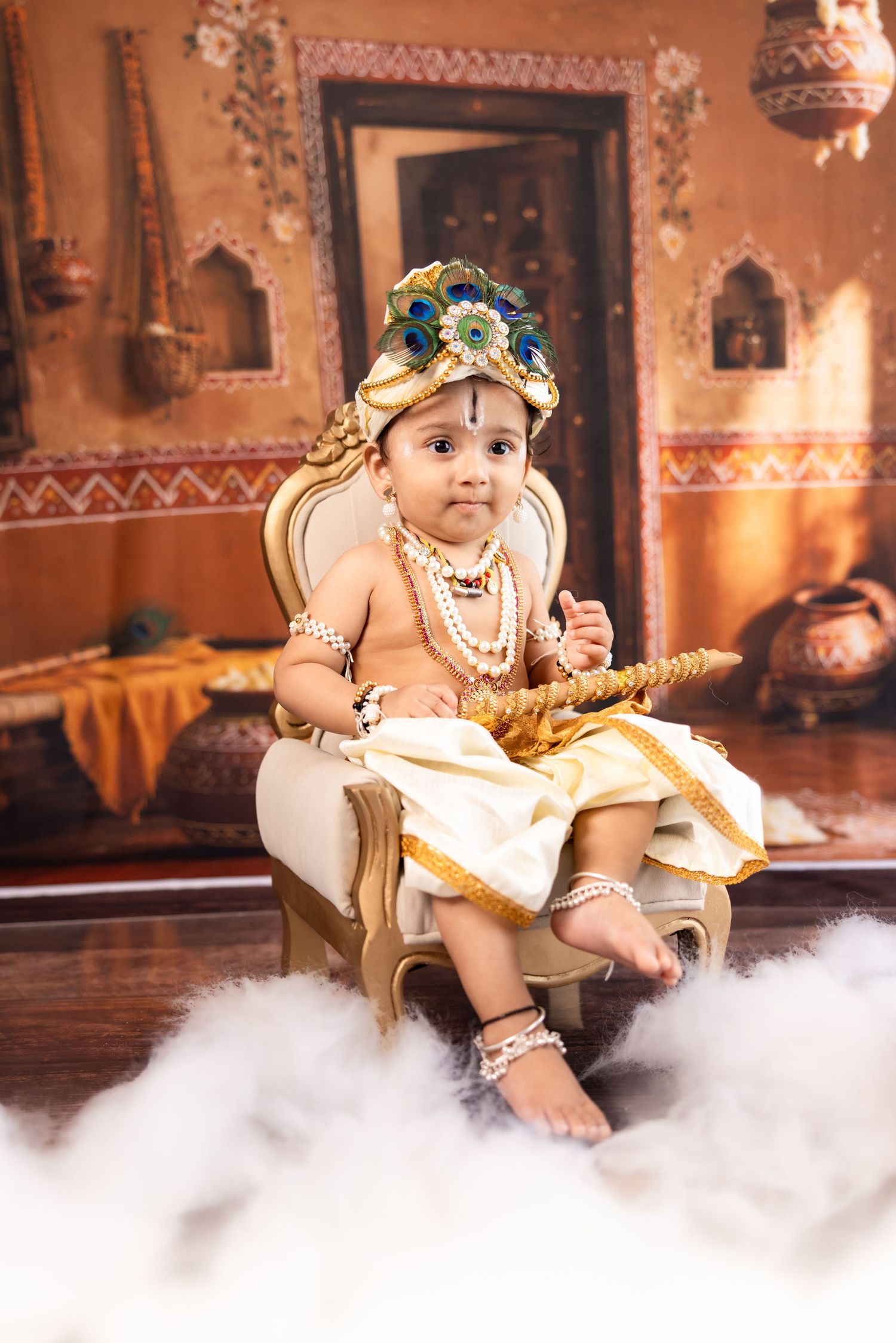 Baby dressed as Lord Krishna in traditional attire with peacock crown, beads, and flute, seated on ornate chair.