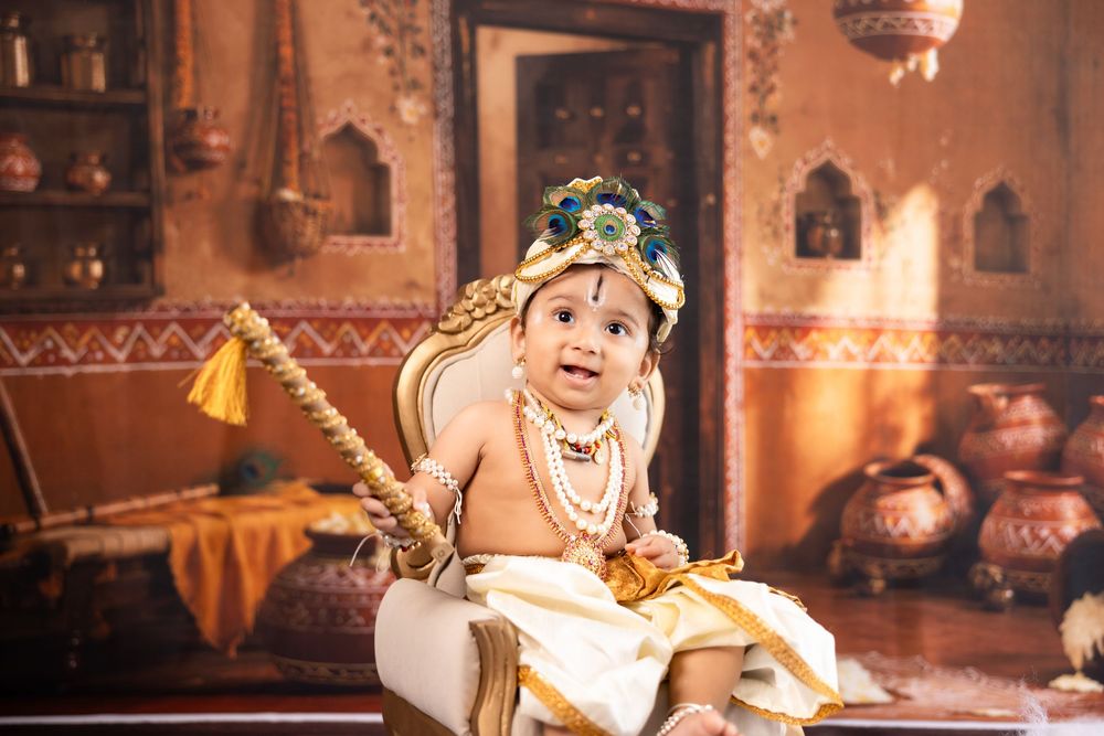 Baby dressed as Lord Krishna in traditional costume with peacock feather crown and gold jewelry in decorative Indian setting.