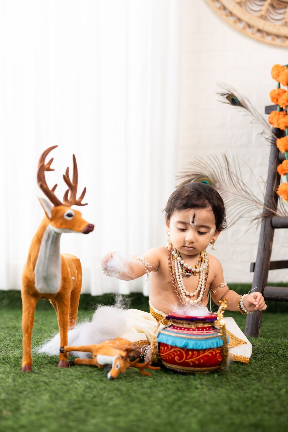 Baby dressed as Lord Krishna playing with decorative pot beside toy deer and peacock feather on green grass.