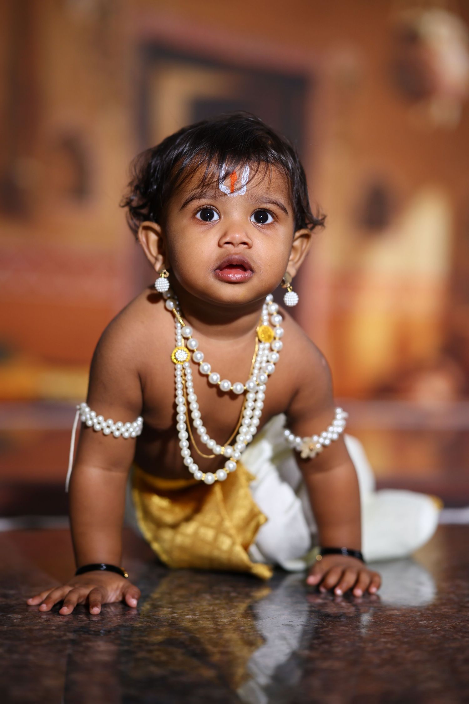 Indian baby dressed as Lord Krishna with pearl jewelry and golden outfit, crawling on floor during festive celebration.