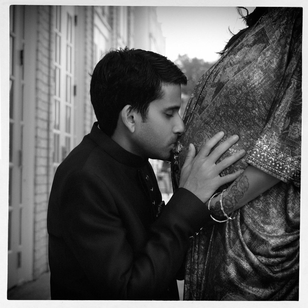 Black and white photo of a man in traditional attire kissing a pregnant woman's belly outdoors.