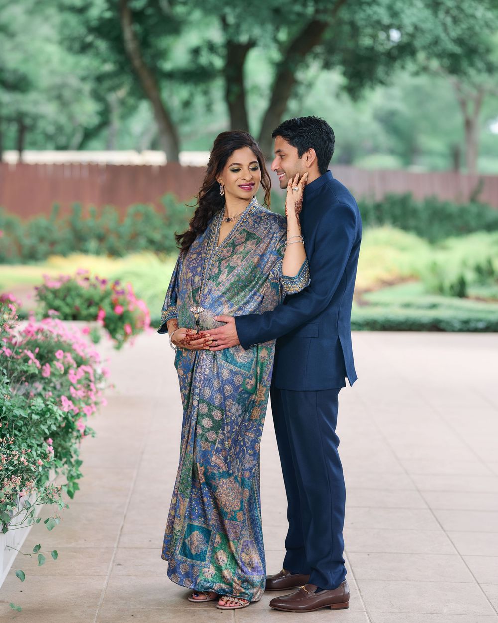 Couple in elegant attire poses in a lush garden, woman in floral sari, man in navy suit whispering softly.