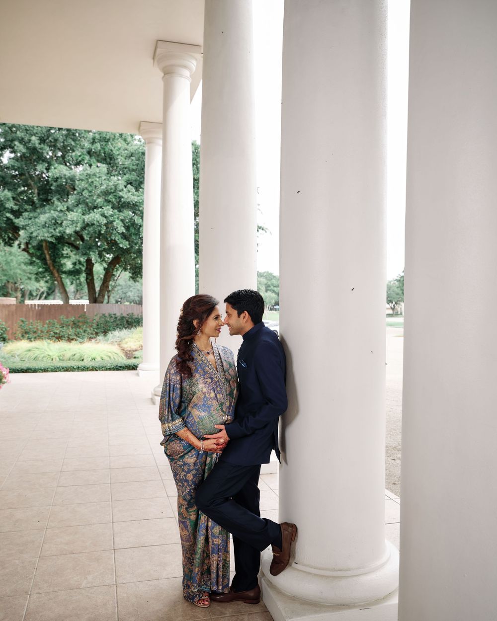 A couple shares a romantic moment leaning against white columns of a classical building surrounded by lush greenery.