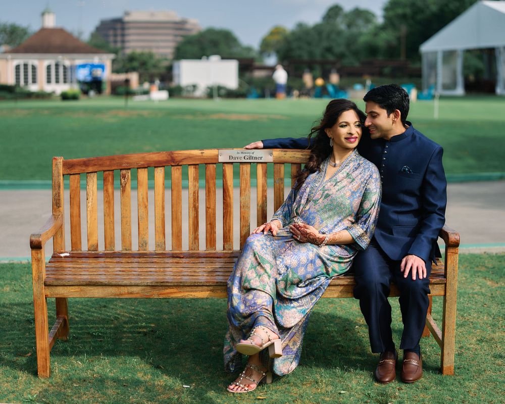 Couple in elegant South Asian attire sitting together on a wooden bench outdoors on a green lawn.