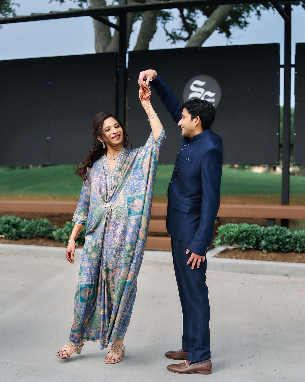 A couple dances outdoors, woman in floral kaftan, man in navy suit, holding hands raised in a spin.