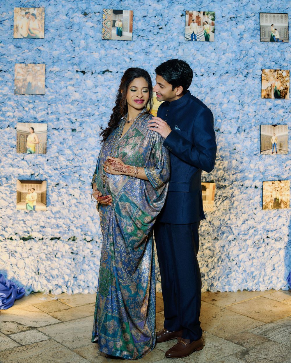 Pregnant woman in blue floral saree with mehndi poses with partner at elegant baby shower with white flower wall backdrop.