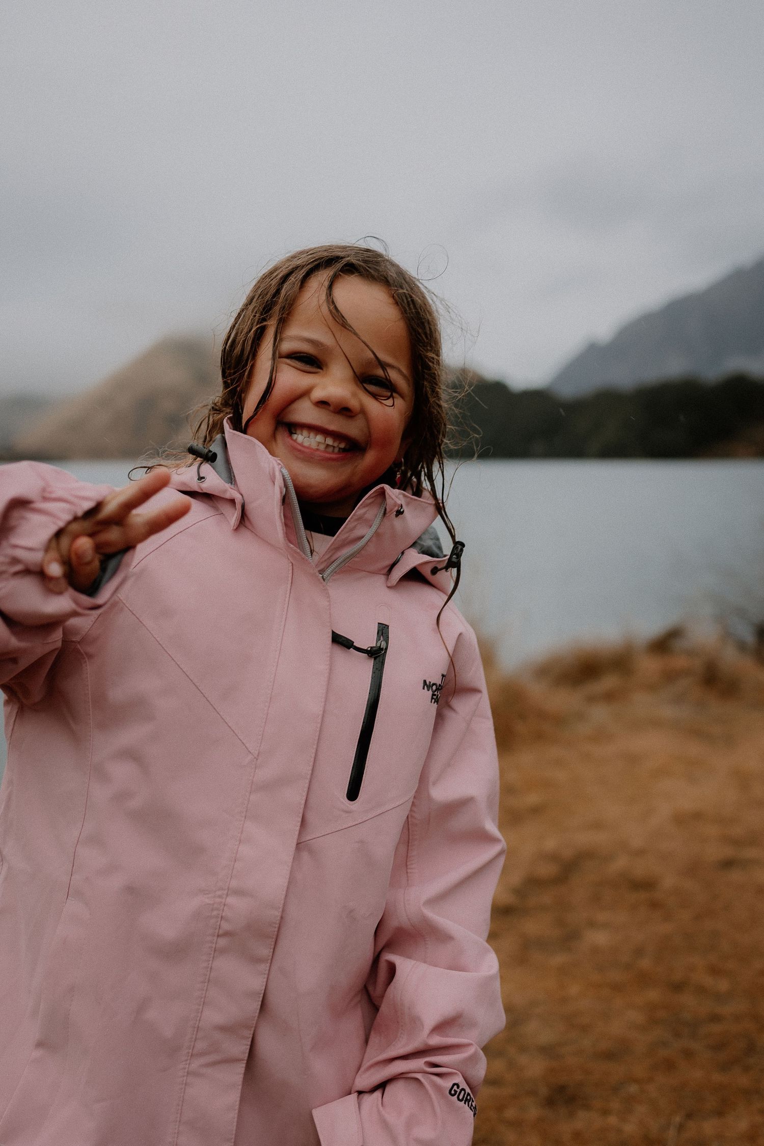 Happy young girl in pink rain jacket making peace sign near mountain lake on overcast day.