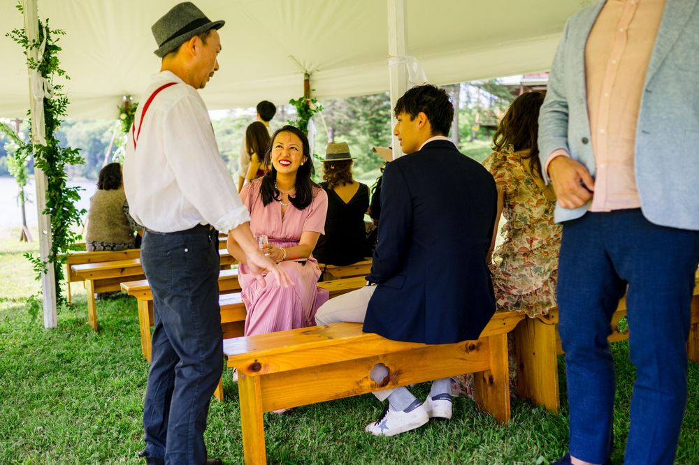 Guests socializing under a white tent at an outdoor wedding reception, seated on wooden benches on green grass.