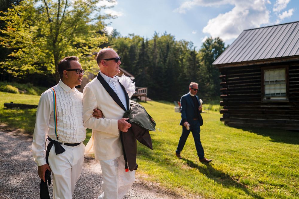 Two men in white suits walk outdoors near a log cabin on a sunny day, with a third man in the background.