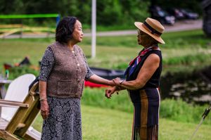 Two women in elegant attire hold hands and converse outdoors near a lush green pond backdrop.